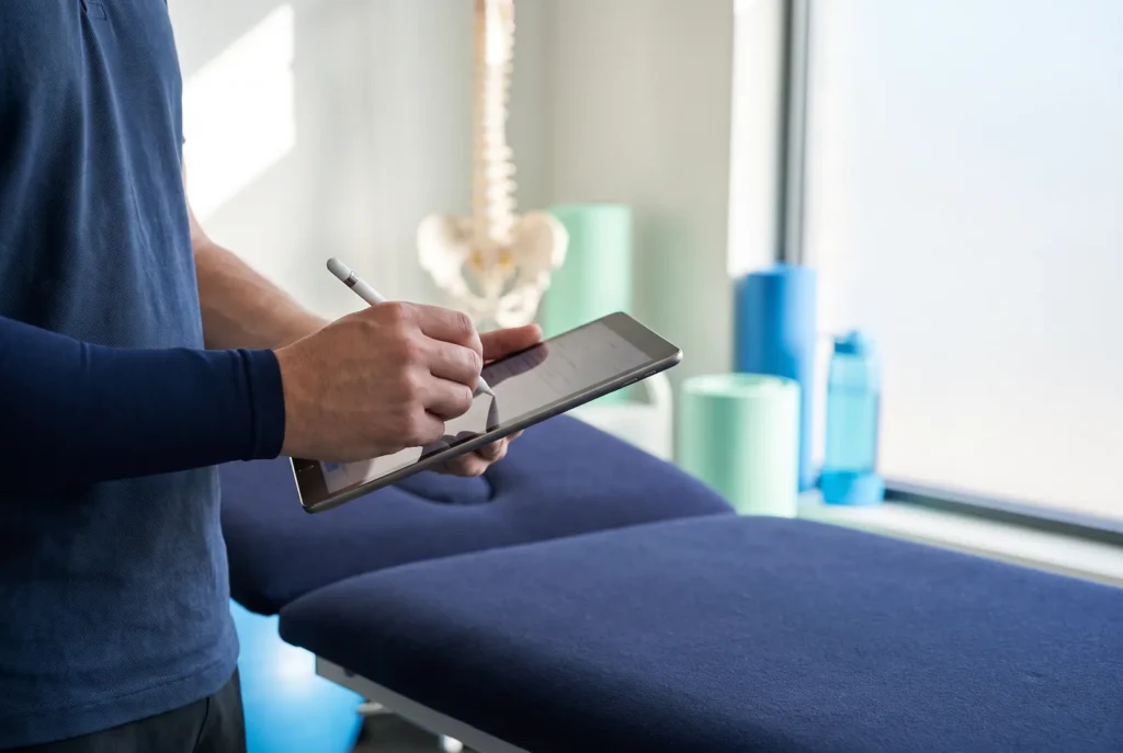 Hände eines Physiotherapeuten schreiben mit Stift auf einem Tablet neben der Behandlungsliege